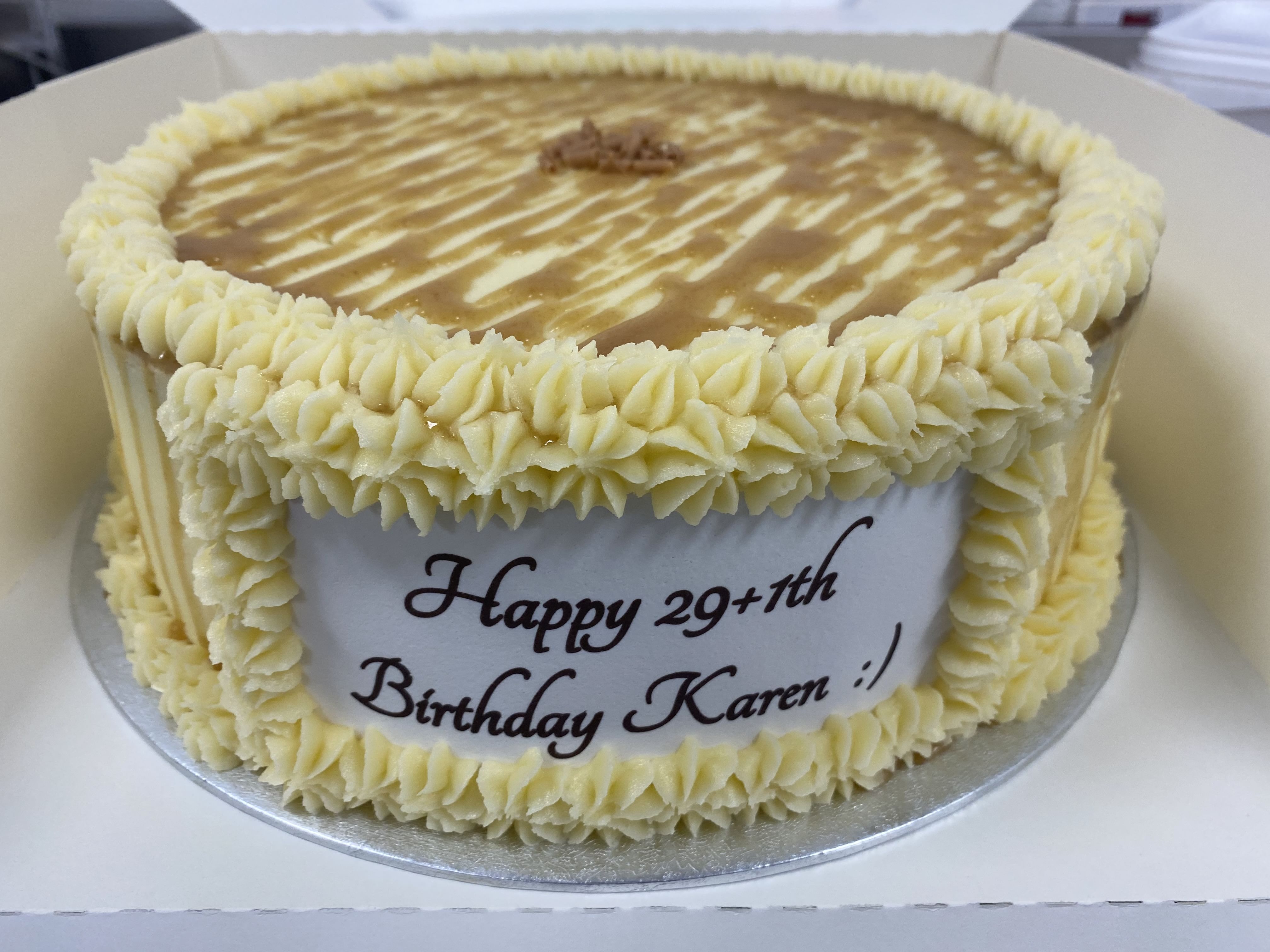 A round Salted Caramel Cake with yellow and tan icing, topped with drizzled homemade salted caramel sauce, and a border of piped rosettes. The side has a white icing plaque with the message 'Happy 29+11th Birthday Karen!' written in brown. The cake is inside a white box.